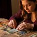 Une jeune femme découvre calmement un ancien jeu de tarot richement illustré sur une table en bois, dans une ambiance chaleureuse aux tons bordeaux et dorés.